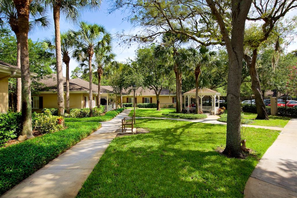 A serene outdoor setting with a pathway winding through lush green grass and trees, surrounded by residential buildings under a clear blue sky.