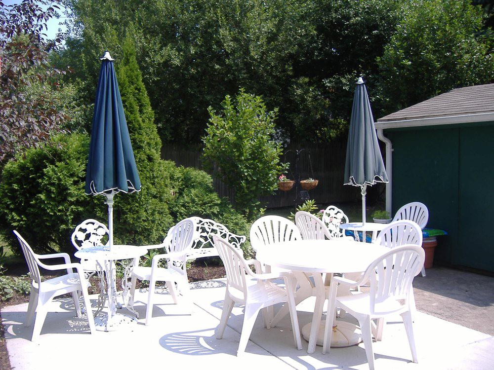 An outdoor patio area with white plastic chairs and tables, shaded by blue umbrellas, surrounded by greenery.