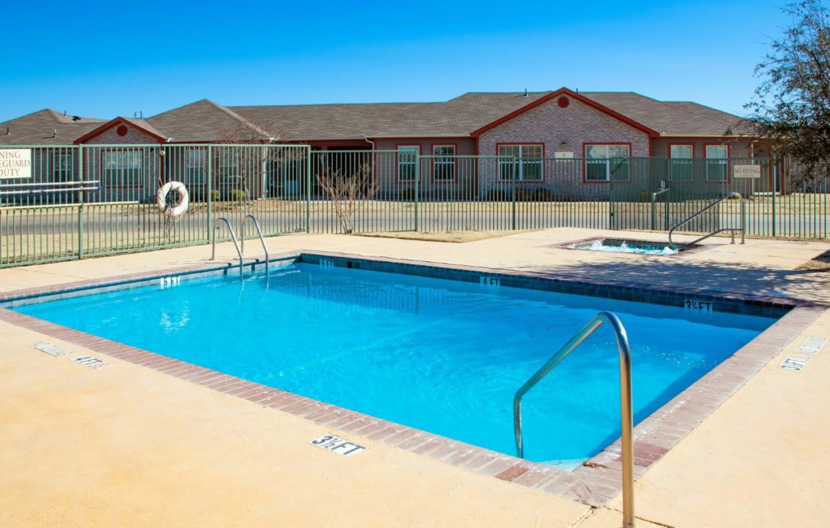 An outdoor swimming pool under a clear blue sky, surrounded by a fence and adjacent to a resort building.