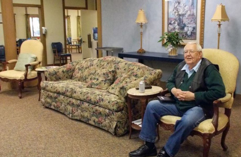 An elderly person sitting in a cozy living room with a floral couch and warm lighting.
