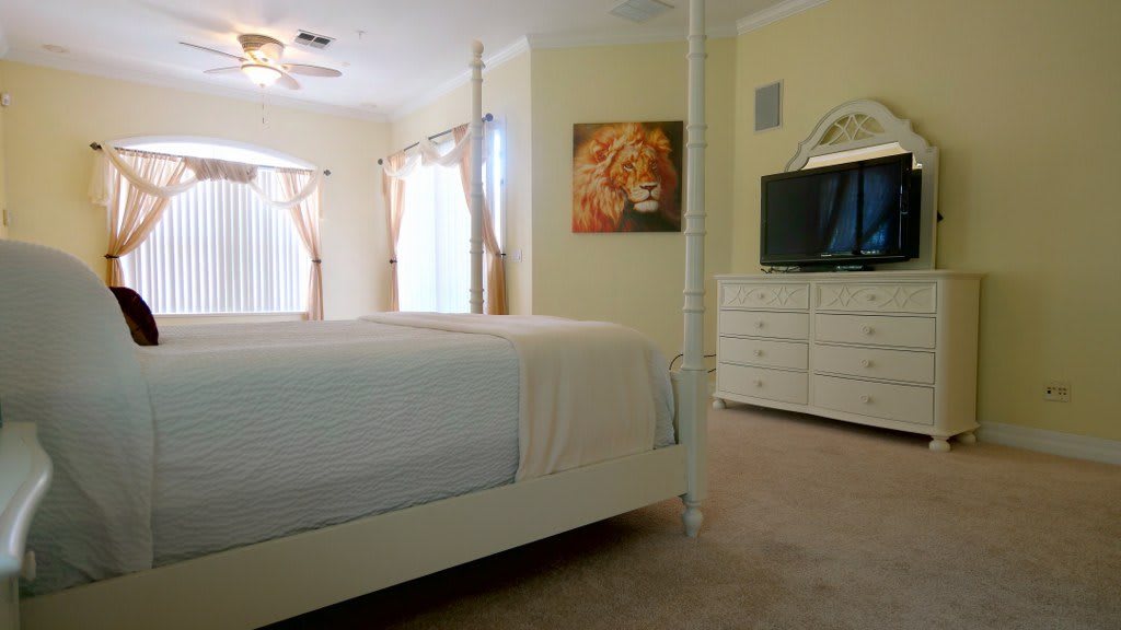 A cozy bedroom with a large bed, dresser, and television, adorned with wall art and natural light streaming through the windows.