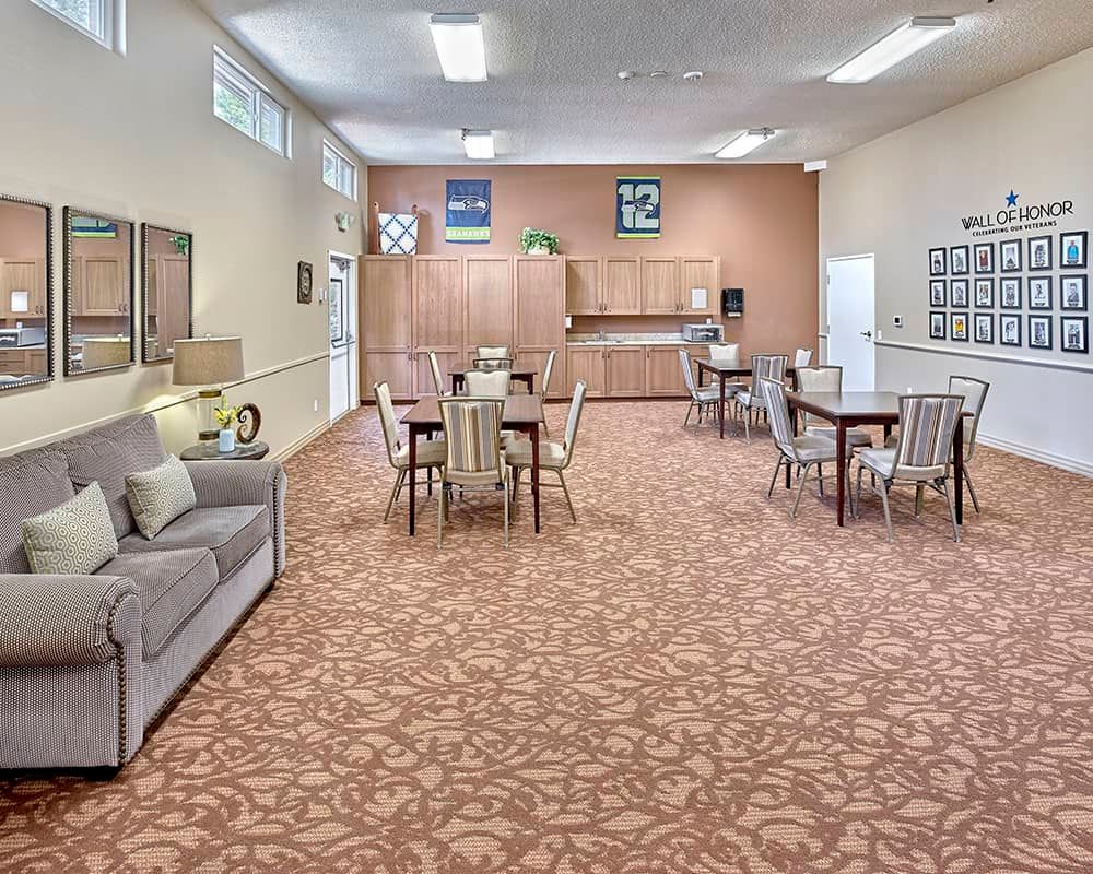 A spacious community room with tables, chairs, and a sofa, featuring a patterned carpet and wall decorations.