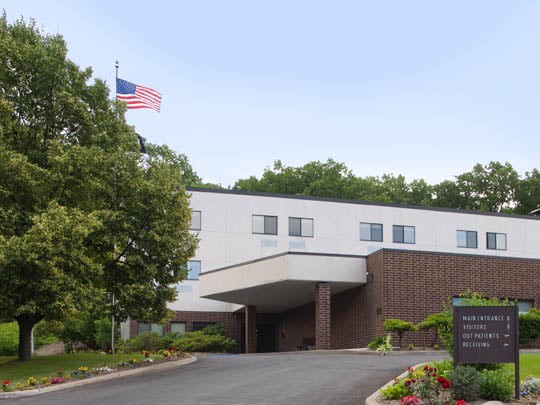 A hotel building with an entrance and driveway, surrounded by trees and landscaping, featuring a flag on a pole.