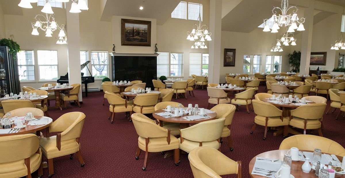 An elegant dining area with multiple tables and chairs arranged on a carpeted floor, illuminated by a chandelier.