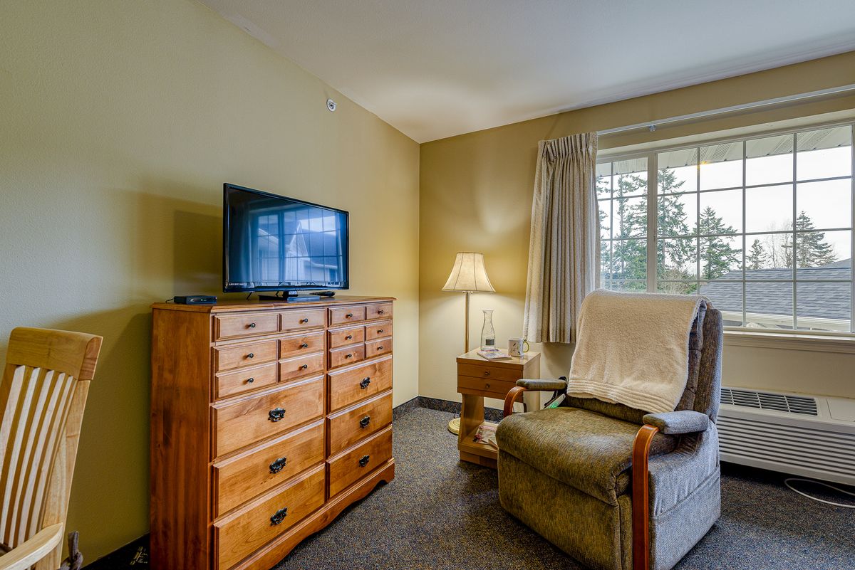 A cozy living room with a television on a wooden dresser, a comfortable chair, and a lamp by the window.