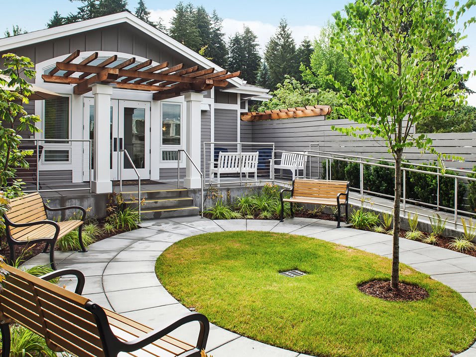 A charming garden space featuring a modern house with a pergola, surrounded by lush greenery and benches.