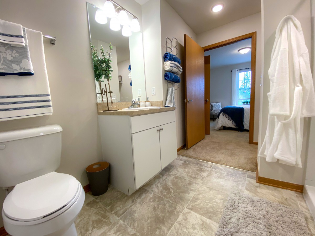 A modern bathroom with a vanity, mirror, and neatly arranged towels leading into a bedroom.