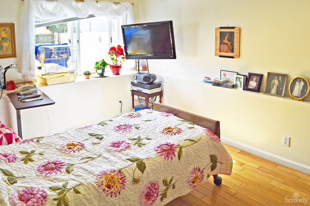 A cozy bedroom featuring a floral bedspread, wall-mounted TV, and decorative frames by the window.