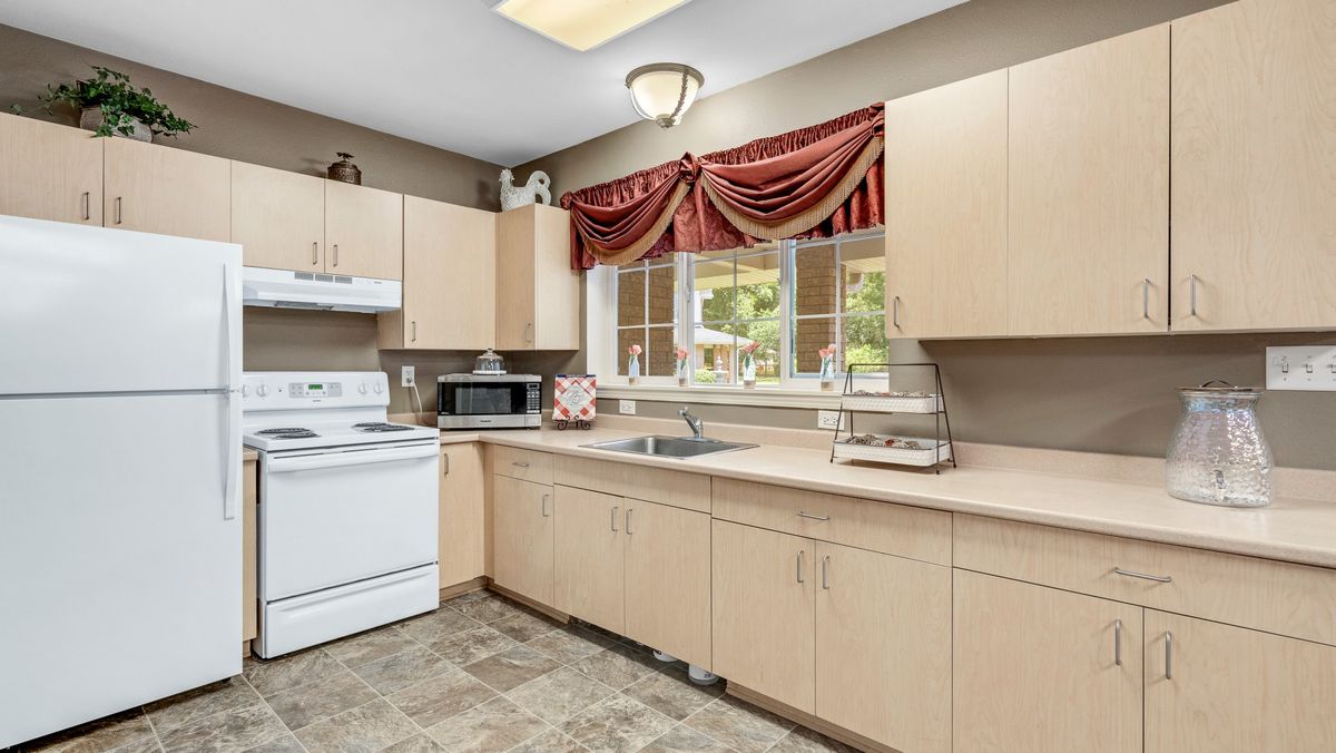 A modern kitchen with light wooden cabinets, a white stove, and a microwave under a large window with red curtains.