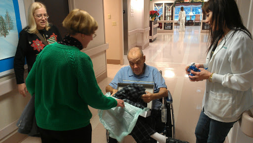 A heartwarming moment of gift-giving at a hospital, where a caregiver presents a surprise to an elderly man in a wheelchair.