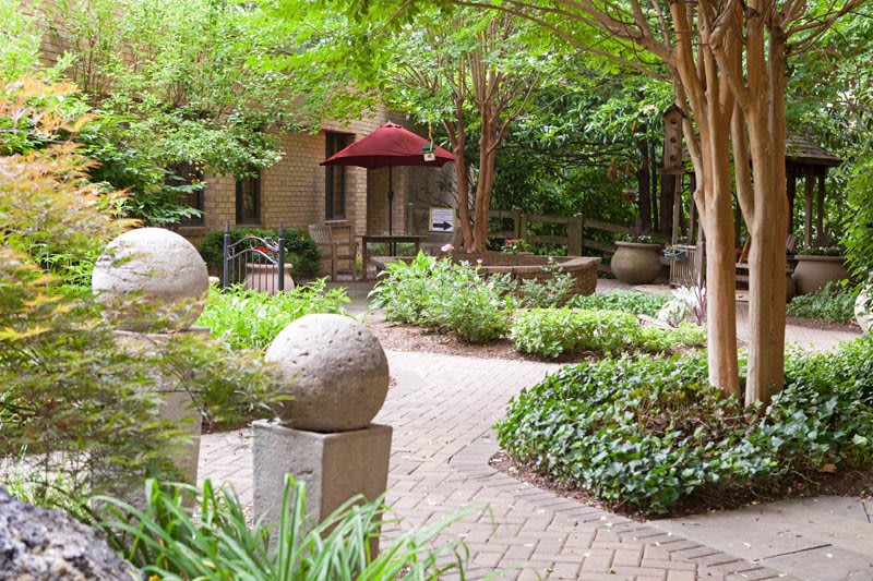 A peaceful garden area with sculptures, greenery, and a winding walkway under trees.