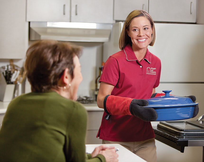 A woman in a red shirt is carrying a blue pot while another person sits at a kitchen counter, engaging in conversation.