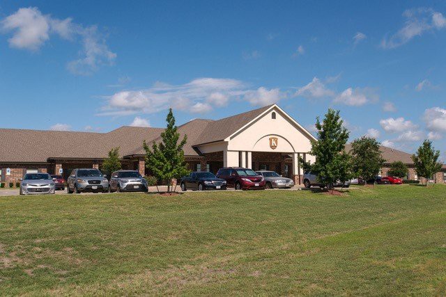 A building with a parking lot surrounded by grass and trees under a partly cloudy sky.