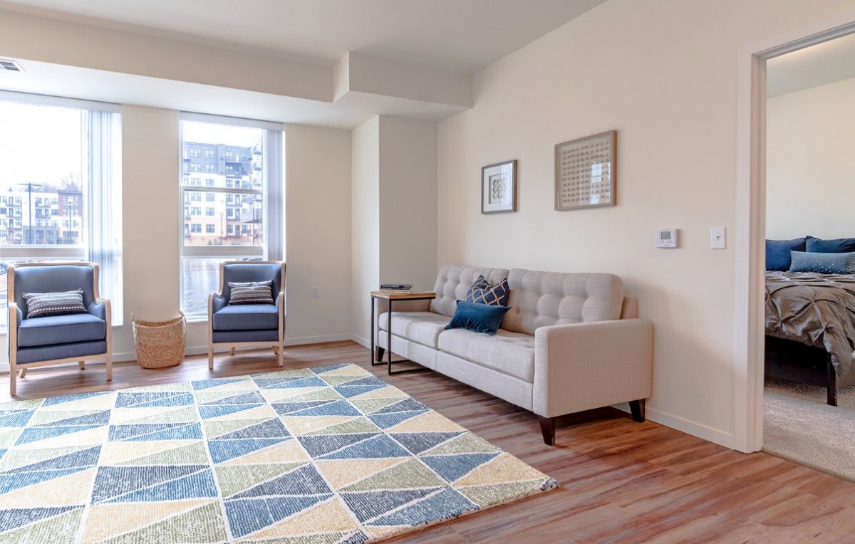 A modern living room with a beige sofa, blue and white rug, and an armchair by the window.
