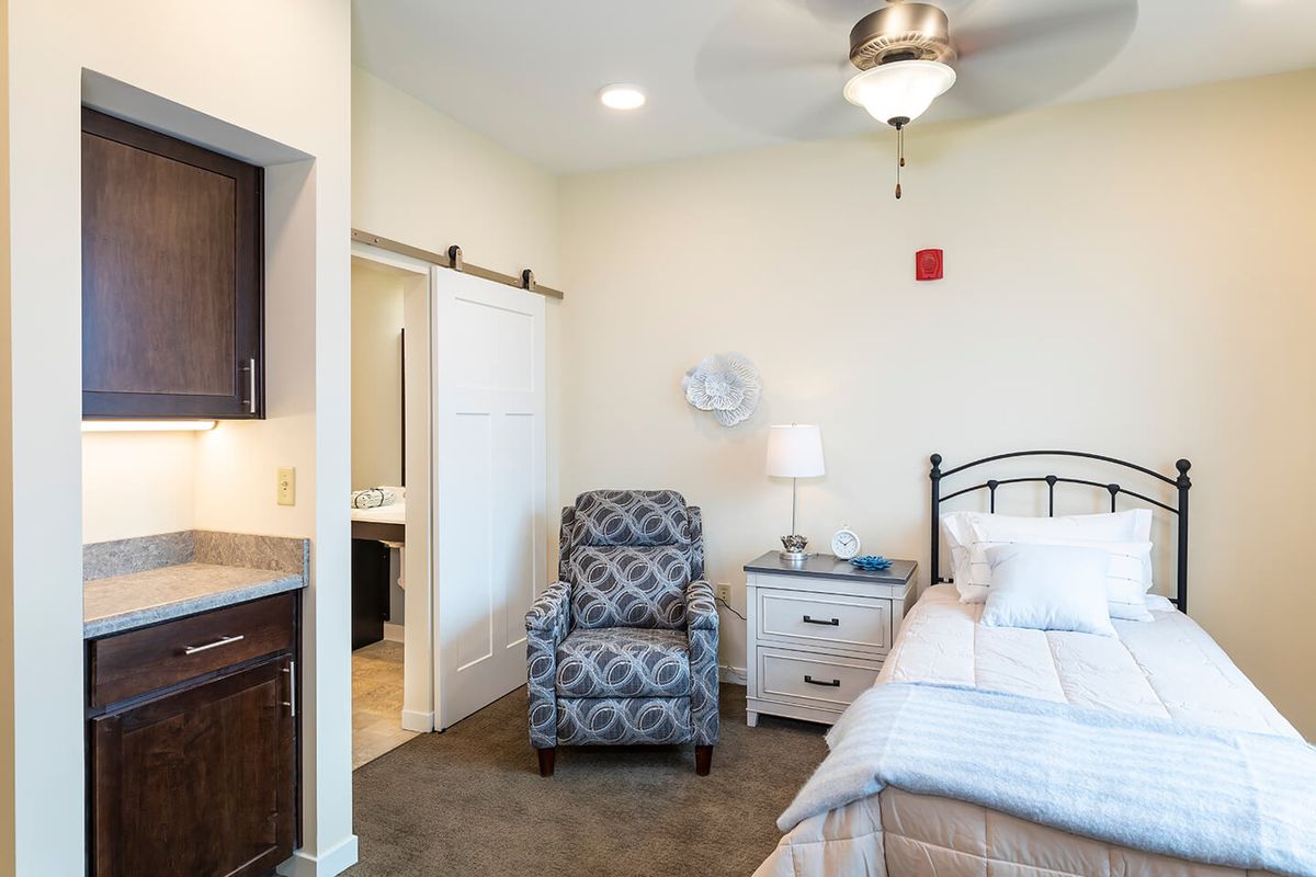 A cozy bedroom with a bed, patterned chair, bedside drawer, lamp, and ceiling fan.