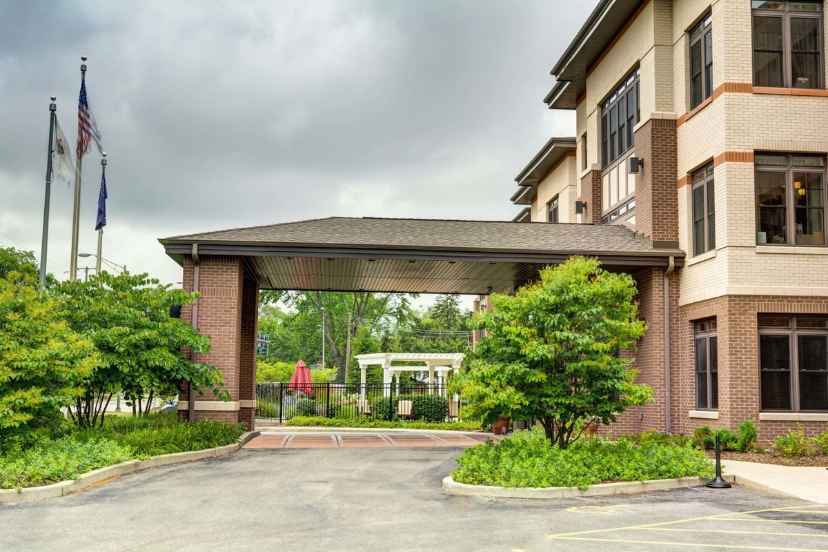 A building entrance with a covered driveway, surrounded by greenery under a cloudy sky.