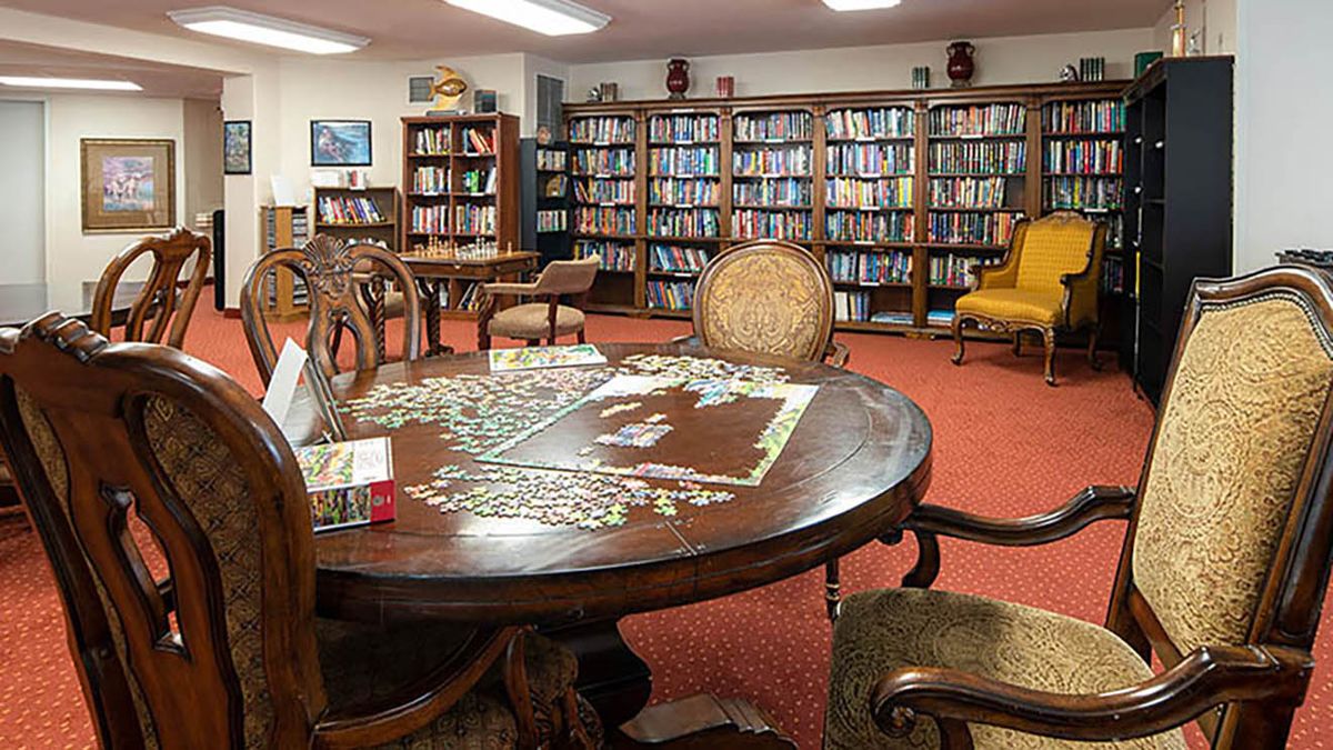 A cozy library room with a table and chairs, featuring bookshelves filled with books and a puzzle in progress on the table.