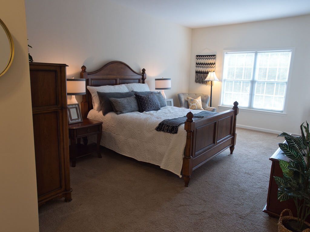 A cozy bedroom with a wooden bed frame and matching furniture, well-lit by lamps and natural light from the window.