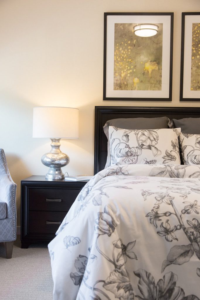 A cozy bedroom with elegant decor, featuring a bed with floral bedding, a nightstand with a lamp, and framed artwork on the wall.