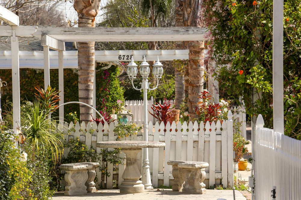 A charming garden featuring a white picket fence, pergola, and outdoor seating surrounded by lush greenery and palm trees.