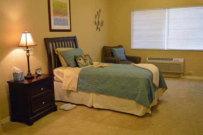 A cozy bedroom with a neatly made bed, cushioned chair, and soft lighting.
