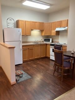 A compact apartment kitchen featuring wooden cabinets, a refrigerator, stove, microwave, and a small dining area with a table and chairs.