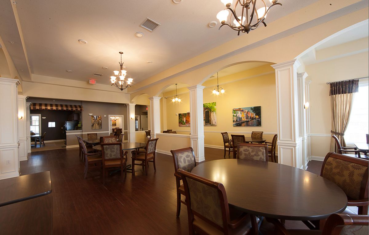 A warmly lit dining area with elegant chandeliers, tables, and chairs arranged on wooden flooring.
