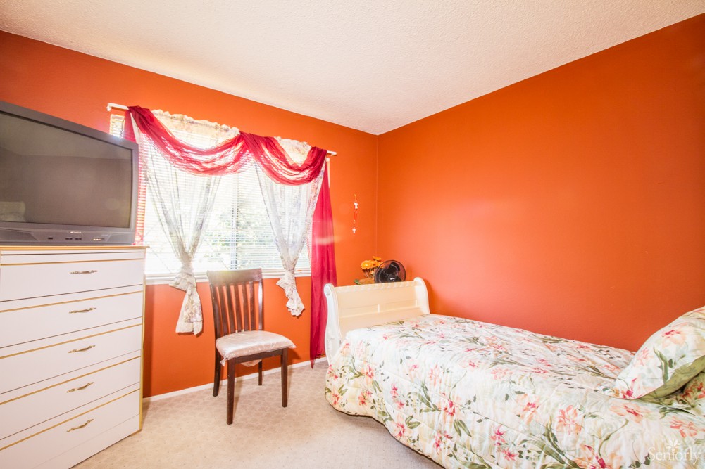 A cozy bedroom with vibrant orange walls, featuring a single bed with floral bedding, a window adorned with curtains, a wooden chair, and a dresser.