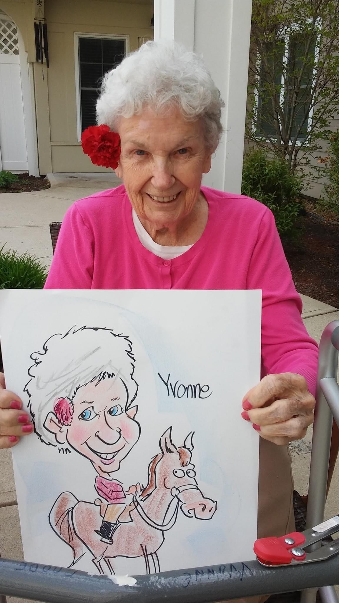 An elderly woman smiling and holding a caricature portrait of herself, wearing a pink shirt with a red flower in her hair.