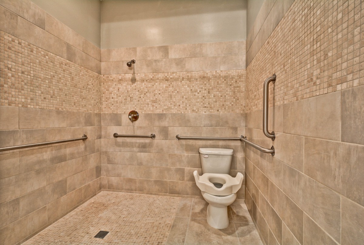 A spacious bathroom with tiled walls, featuring a toilet and shower area equipped with handrails for accessibility.