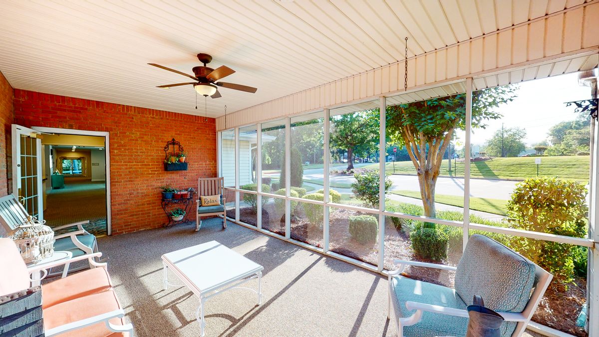 A cozy screened-in porch featuring a ceiling fan, rocking chair, and coffee table with large windows overlooking a garden.