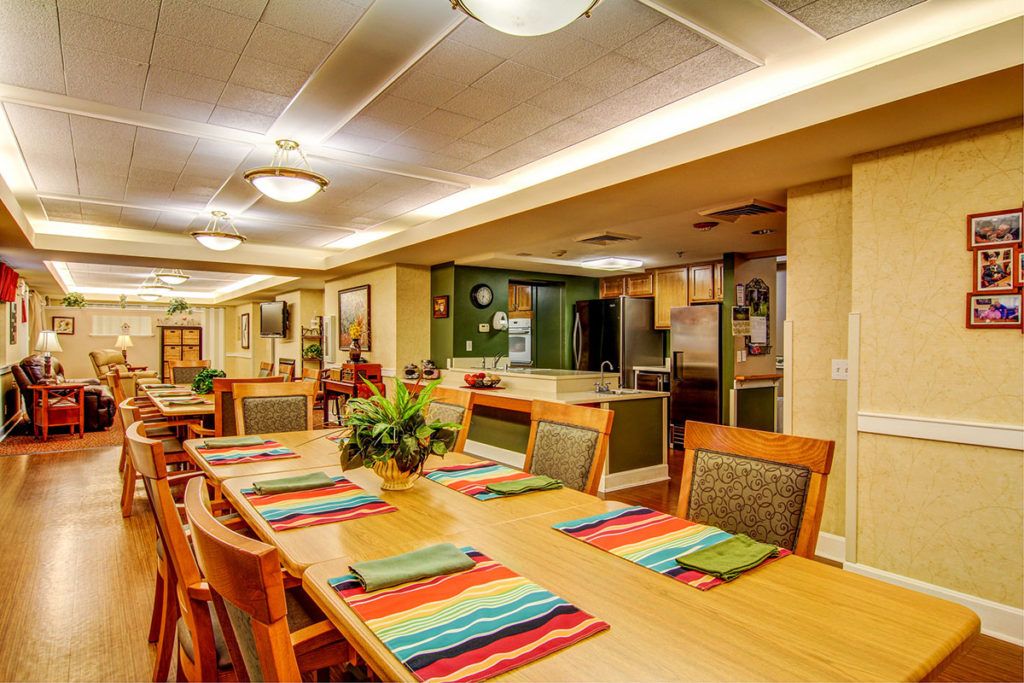 A warmly lit dining area with a long wooden table and colorful placemats, adjacent to an open kitchen.