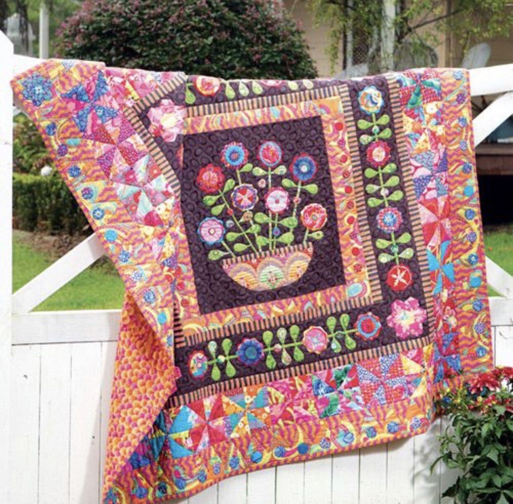 A colorful quilt featuring a floral design is hung outdoors on a white fence.