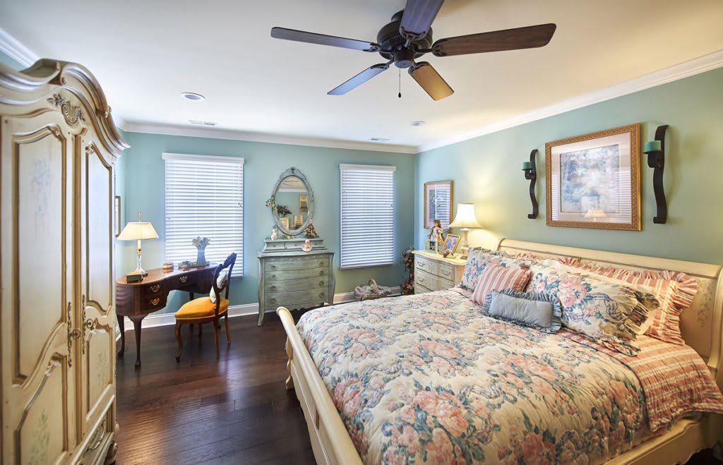 A cozy bedroom with floral bedding and elegant furniture, featuring a ceiling fan and wooden floors.