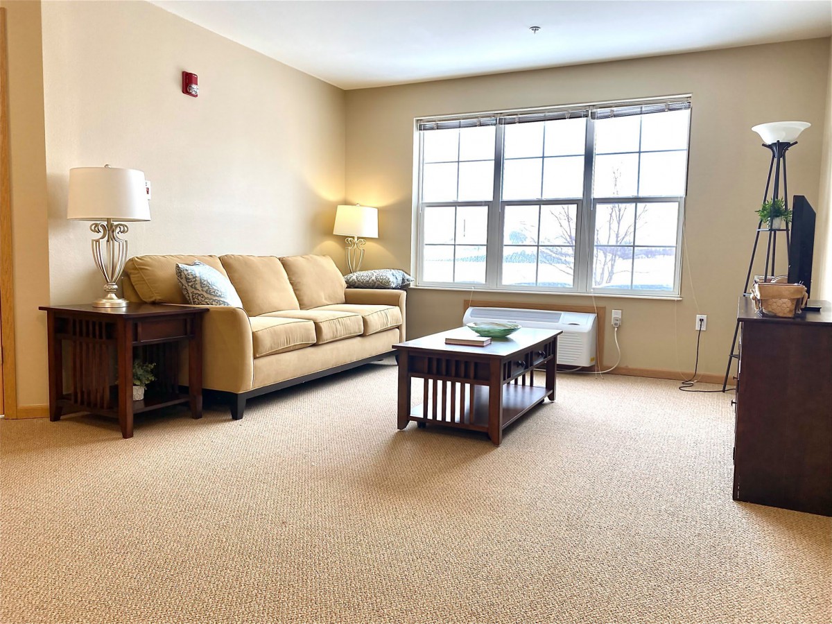 A cozy living room with a beige sofa, wooden coffee table, and a large window letting in natural light.