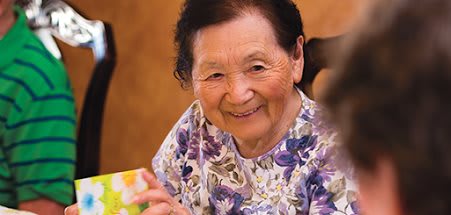An elderly woman smiling warmly indoors, wearing a floral shirt.