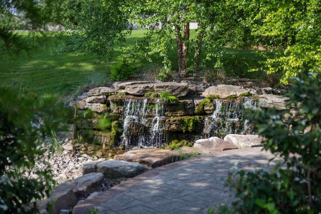 A tranquil waterfall cascading over rocks surrounded by lush greenery and a stone pathway.