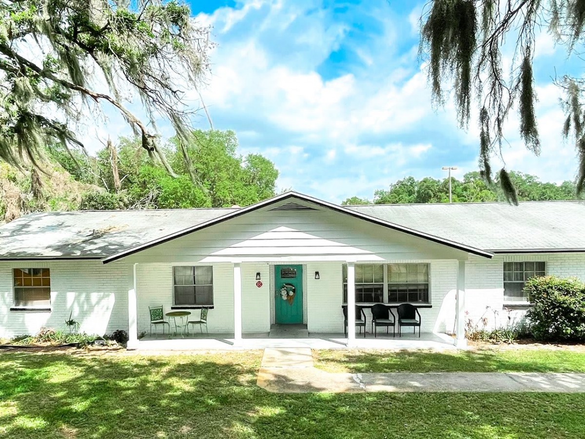 A cozy single-story house with a white exterior and a small front porch, surrounded by lush greenery and under a clear blue sky.
