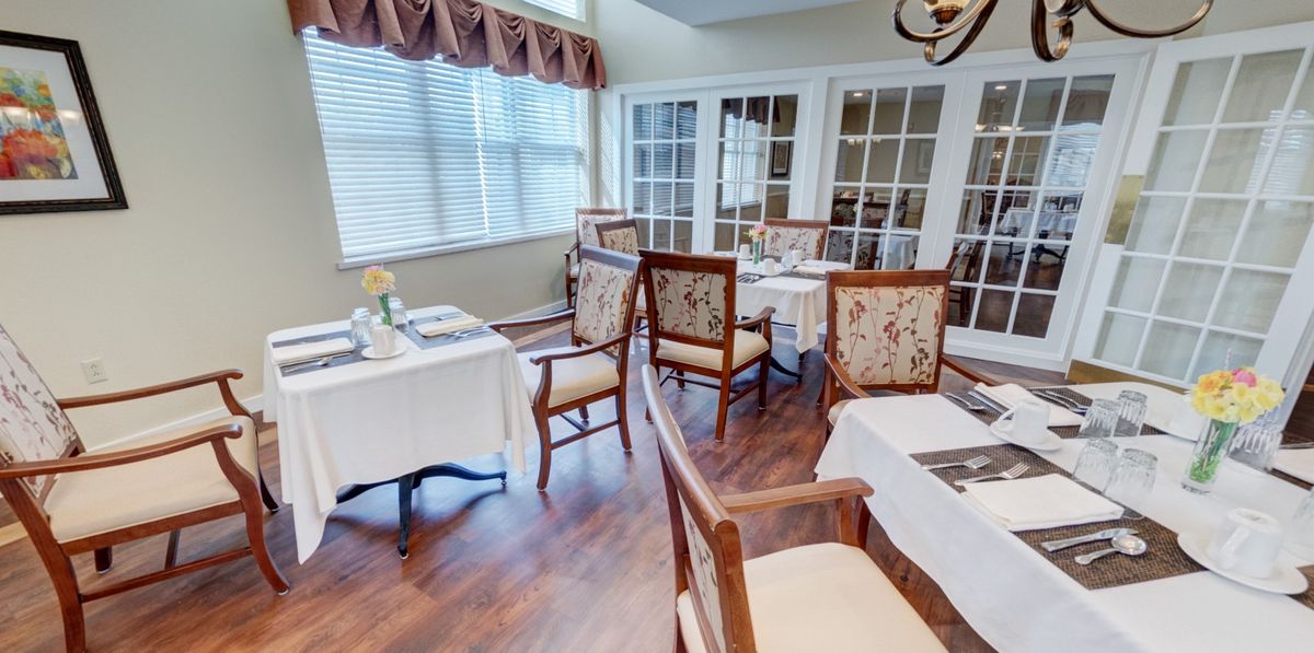 A cozy dining room with elegantly set tables and floral decorations, featuring wood flooring and abundant natural light.