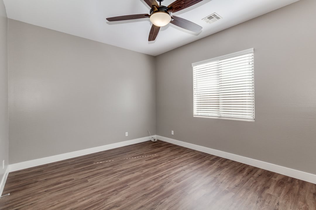 An empty room with a ceiling fan, wood flooring, and a window with blinds.