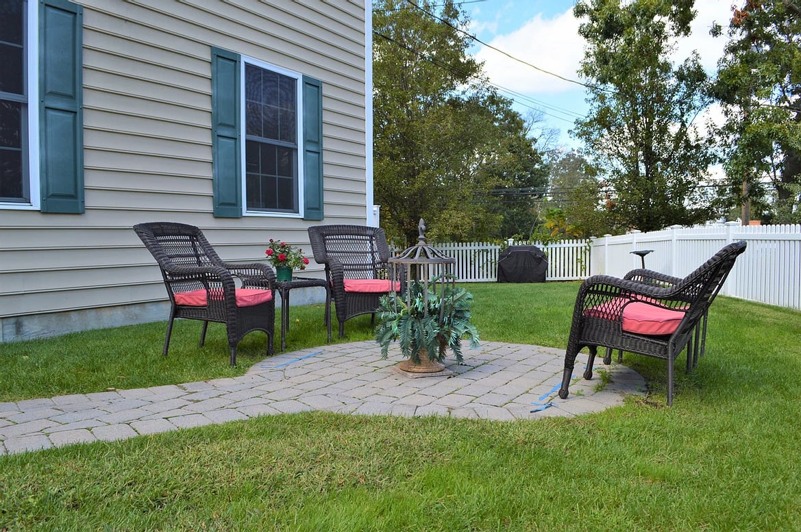 A cozy backyard patio area with wicker chairs and a small table on a paved surface, surrounded by a lawn.