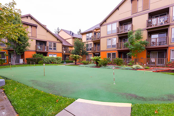 A residential apartment complex featuring a well-maintained putting green area surrounded by landscaped greenery.