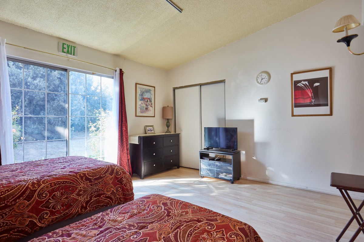 A cozy bedroom with two beds, a dresser, and a television, featuring natural light from a large sliding door.