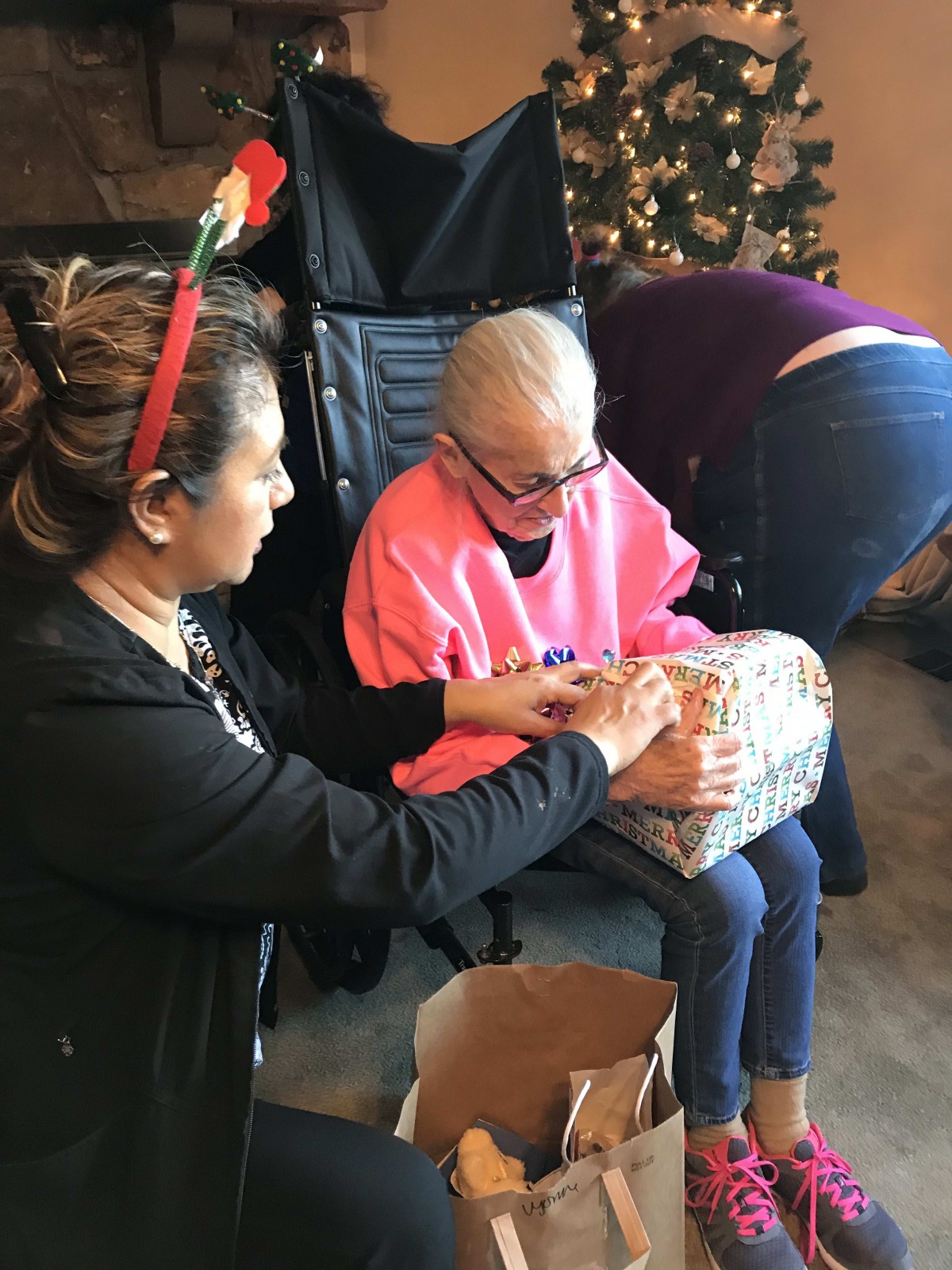A family gathers to share Christmas cheer, as one person helps an elderly woman open a gift beside a decorated tree.