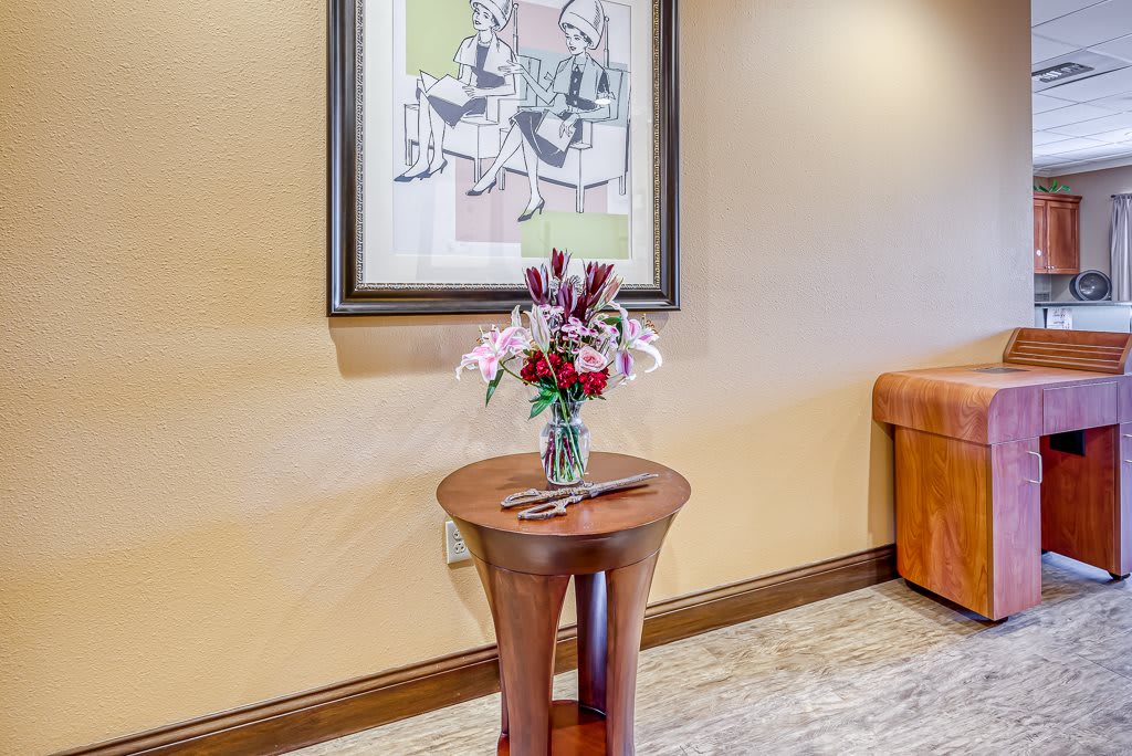 A small wooden table with a vase of flowers is placed below a framed artwork in a well-lit room.