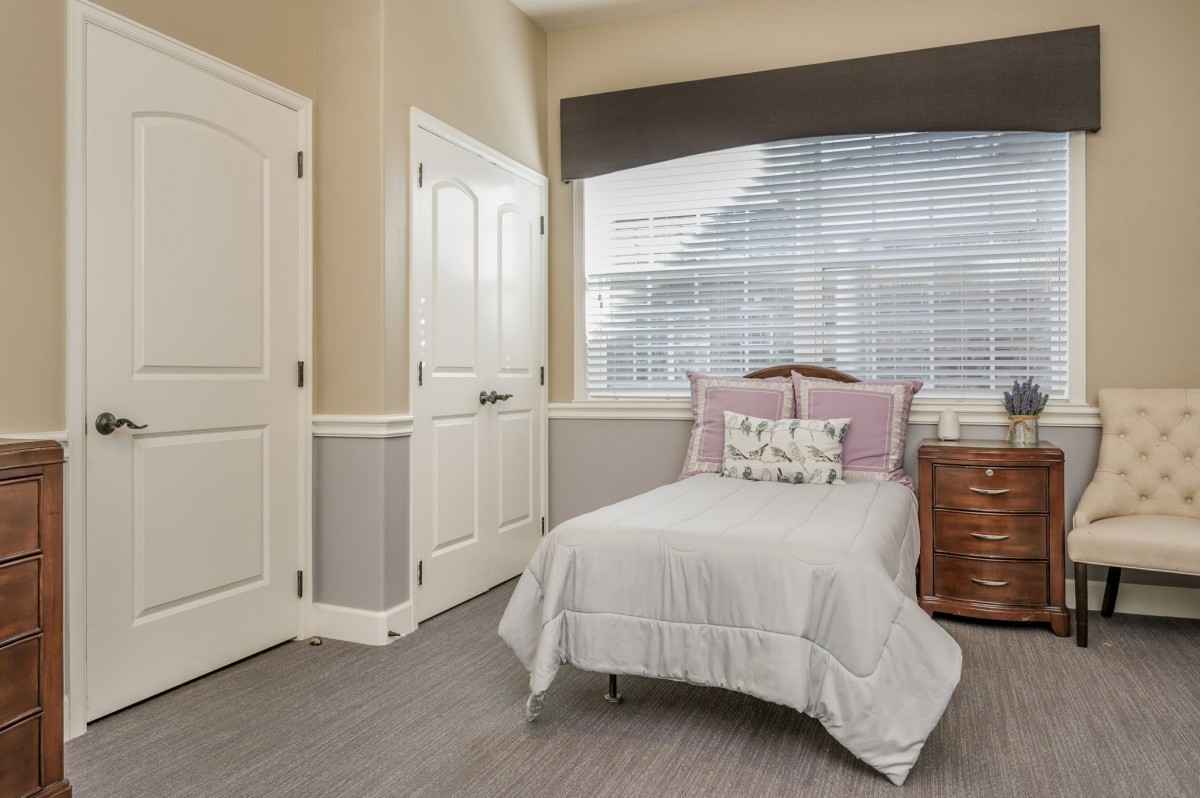A cozy bedroom featuring a single bed with decorative pillows, a wooden bedside table, and a window with blinds.