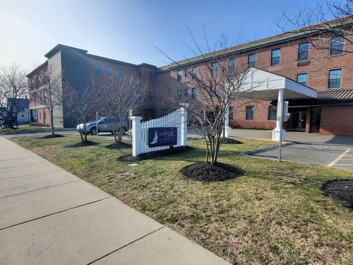 A sunny view of a building with a sign on the front lawn and parked cars nearby.