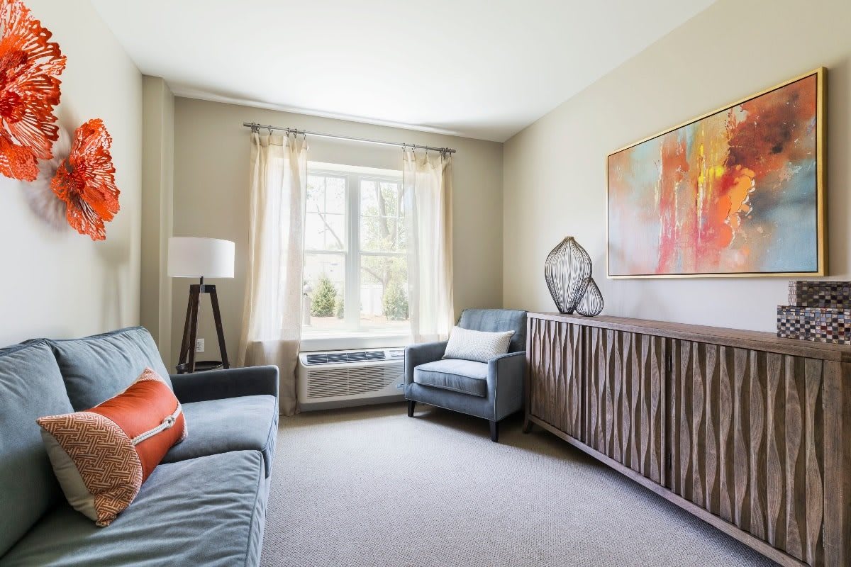 A cozy living room with modern furnishings, including a sofa and armchair, accented by a large window and colorful artwork.