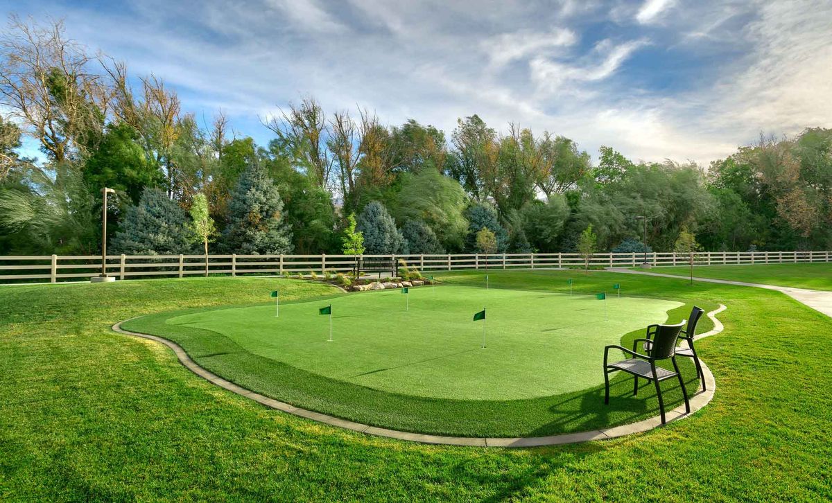 A scenic outdoor putting green surrounded by lush trees and a white fence, with a bench nearby.