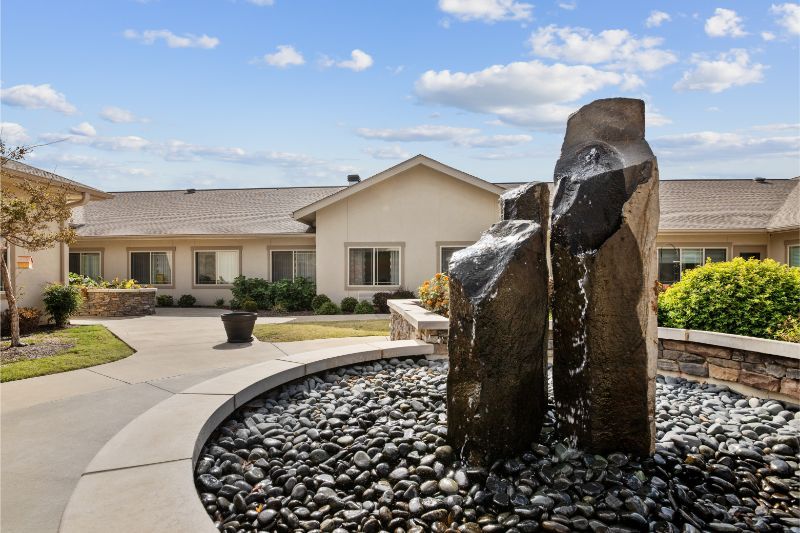 A modern outdoor fountain with stone features enhances the landscape in front of a house under a clear blue sky.